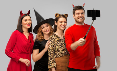 Poster - friendship, holiday and people concept - group of happy smiling friends in halloween costumes of witch, devil and cheetah taking picture by smartphone on selfie stick over grey background