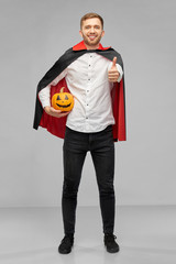 Poster - holiday, theme party and people concept - happy smiling man in halloween costume of vampire with dracula cape showing thumbs up and holding jack-o-lantern pumpkin over grey background