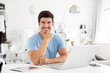 © BestForYou - Smiling man in a blue t-shirt sitting at office desk behind laptop. Cheerful freelance.