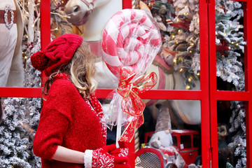 Wall Mural - Girl in a red sweater with lollipop enjoys the magic of Christmas holidays standing on a street at a toy store window