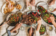 Summer barbeque party. Flat-lay of table with grilled meat, vegetables, salad, roasted potato and peoples hands with cutlery over white tablecloth, top view. Family gathering, comfort food concept