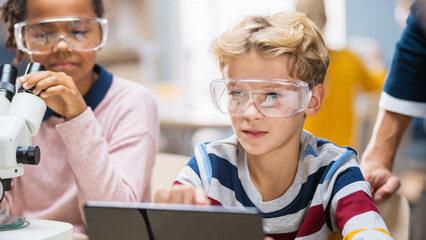 Poster - Elementary School Science Classroom: Boy Uses Digital Tablet Computer to Check Information on the Internet while Enthusiastic Cute Little Girl Uses Microscope.