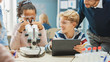 Elementary School Science Classroom: Cute Little Girl Looks Under Microscope, Boy Uses Digital Tablet Computer to Check Information on the Internet, while Enthusiastic Teacher Explains Lesson