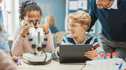 Poster - Elementary School Science Classroom: Cute Little Girl Looks Under Microscope, Boy Uses Digital Tablet Computer to Check Information on the Internet, while Enthusiastic Teacher Explains Lesson