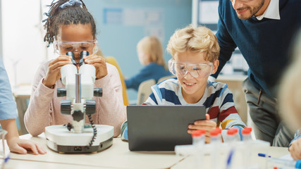 Poster - Elementary School Science Classroom: Cute Little Girl Looks Under Microscope, Boy Uses Digital Tablet Computer to Check Information on the Internet, while Enthusiastic Teacher Explains Lesson
