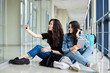 © Natalia - Two young brunette girls, sitting on floor in light airport hallway, with luggage behind, taking selfie with phone, smiling, wearing casual clothes. Girlfriends, traveling by air, waiting for flight.