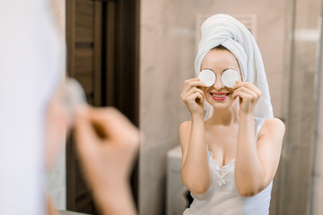 Young beautiful smiling woman with towel on her head looking in the mirror in bathroom, laughing and having fun, while hiding her eyes with two cotton pads. Morning skincare routine