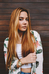 Portrait of casually-dressed female hipster standing on wooden wall background looking to you, young trendy woman posing outdoors enjoying good day in summer