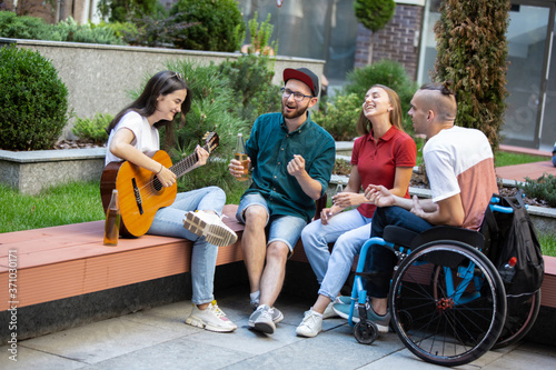 Drinks and songs. Group of friends taking a stroll on city\'s street in summer day. Handicapped man with friends having fun. Inclusion and diversity concept, normal lifestyle of special groups of