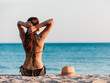 © Vitalie - The young girl sitting on the sand on the beach, looking at the sea water and squeezing her brunette hair. The hat sits next to the girl on the sand. Girl in a bathing suit relaxing at the ocean
