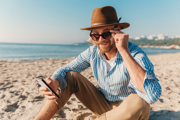 young attractive hipster man sitting on beach by sea on summer vacation, boho style outfit, holding using internet on smartphone, dressed in shirt, sunglasses and hat