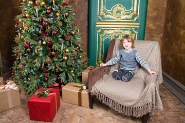 wearing in grey jeans and sweater happy man-child sits in armchair near new year tree and presents