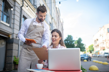 Brown-haired female sitting at table, holding menu, consulting with bearded waiter