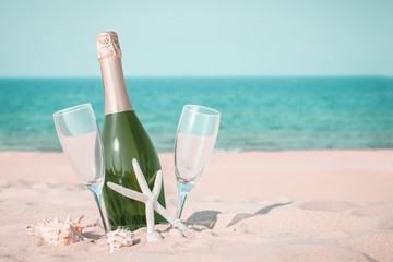 A bottle of champagne and two glasses on a sandy beach with seashells against a blue sea background