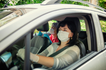 Caregiver and an elderly woman wearing protective masks drive a car for regular medical appointments. The carer's face is out of focus, the old woman is clearly visible.