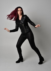 Wall Mural - Full length portrait of  girl with red hair wearing black leather jacket, pants and boots. Standing pose, isolated against a grey studio background.