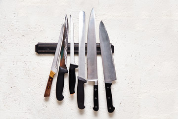 A set of large old chef's knives on a magnetic holder hang on a white wall. Food preparation concept, kitchen attributes, old worn-out knives.