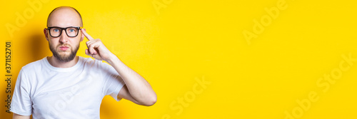 Fotografía Serious bearded bald young guy in glasses holds a finger to his temple on a yellow background