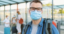 Portrait Of Handsome Caucasian Man In Glasses And Medical Mask Looking Behind, Then At Camera And Standing At Bus Stop Or Train Station. Wife With Kids And Suitcases On Blurred Background. Pandemic.