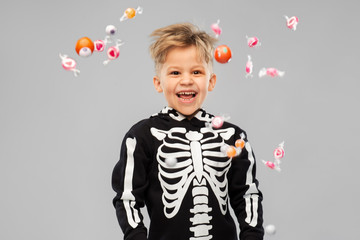 halloween, holiday and childhood concept - smiling boy in black costume of skeleton with candies trick-or-treating over grey background