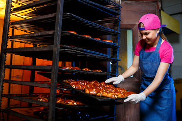 Young girl baker holds a tray with hot pastries against the background of an industrial oven in a bakery. Production of bakery products. Fresh crispy pastry rack