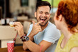 Cheerful man laughing while eating donut and talking to his girlfriend in a cafe.