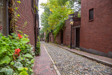 Narrow Cobbled Alley Lined With Old Brick Residential  Buildings And Gas Lit Lamp Posts On A Cloudy Autumn Day