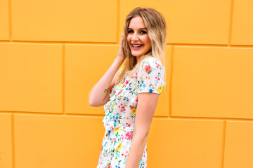 Wall Mural - Bright sunny portrait of pretty blonde woman wearing floral sexy dress, posing near yellow wall, making surprising face, toned colors.