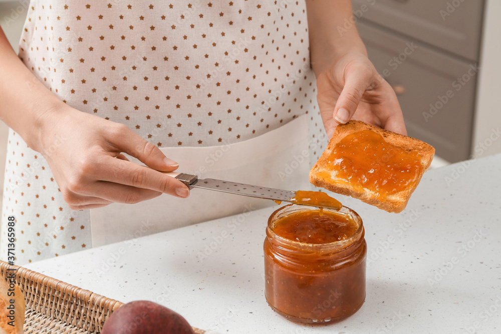 Woman spreading tasty peach jam on bread slice in kitchen
