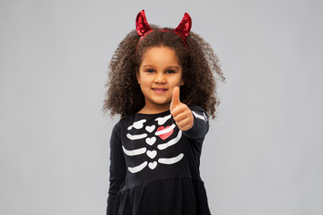 Wall Mural - halloween, holiday and childhood concept - smiling african american girl in black costume dress and red devil's horns over grey background