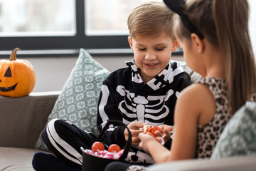 Wall Mural - halloween, holiday and childhood concept - smiling little boy and girl in party costumes with candies sitting on sofa at home