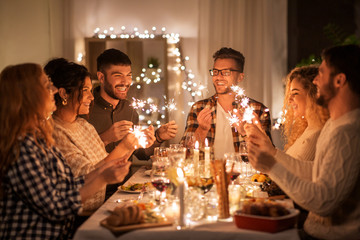 holidays, celebration and people concept - happy friends with sparklers having christmas dinner at h