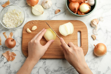 Woman cutting fresh onion on wooden board at white marble table, top view