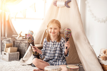 Wall Mural - Happy little girl playing on wooden maracas in room decorated with ethnic drums and wigwam