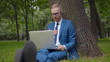© TommyStockProject - Freelancer in headset working in park under tree having video call with client on laptop