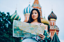 Asian Female Tourist Pondering While Reading Map Choosing Right Direction For Strolling, Surprised Young Girl Get Lost In Moscow City Checking Routes Standing On St. Basil's Cathedral Background