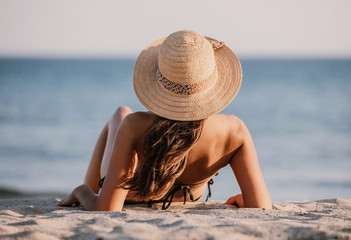 Young lady in a bathing suit and hat sits on the sand on the beach, looking towards the sea. Beautiful girl is facing the ocean with her back to the lens