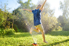Funny Little Boy Playing With Garden Sprinkler In Sunny Backyard. Preschooler Child Laughing, Jumping And Having Fun With Spray Of Water.