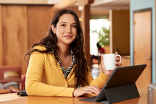 Portrait Of Young Ethnic Woman Wearing Yellow Sweater With Black And White Striped Blouse, Sitting At Bar In Kitchen Of Downtown Loft With IPad And Coffee Mug