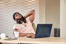 Portrait Young Ethnic Man Sitting At Desk With Laptop Computer Wearing Large Over Ear Headphones Around His Neck 