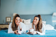 Smiling Town Sisters Holding Container While Lying On Bed In Hotel Room