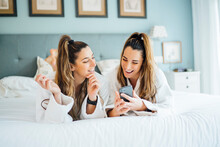 Smiling Twin Sisters Using Smart Phone While Lying On Bed In Hotel