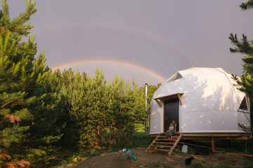 Wall Mural - Mom with kid sitting near geodome. black cat and a dog walking nearby, small bicycle, a pine forest and  beautiful semicircular rainbow in the sky after rain