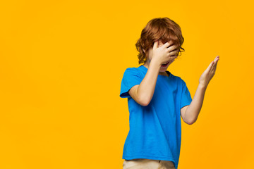  child in a blue T-shirt on a yellow background and red hair touch his face with his hands