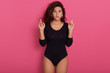 © sementsova321 - Young woman with serious facial expression posing over rosy wall with fingers crossing and wishing best, lady with wavy hair wearing black stylish bodysuit,
