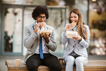 Smiling businessman and businesswoman with sandwiches sitting in front of the office building - lunch break