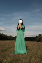 Young Woman In A Field In Green Dress Holding Round Mirror In Front Of Face Reflecting The Sky