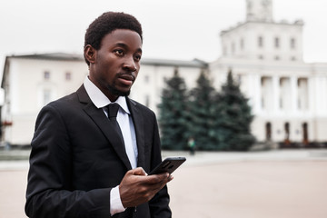 Wall Mural - Selective focus of serious young african american businessman standing outside with phone and looking straight ahead. Business concept with copy space. 