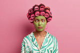 Fototapeta  - Funny ethnic young woman makes grimace, crosses eyes, applies hair rollers, makes hairstyle for special day, wears green moisturizing mask on face, dressed in domestic clothes, poses in studio
