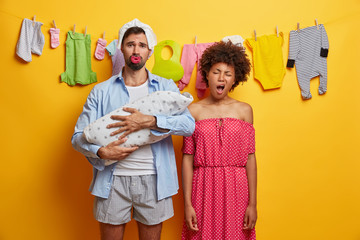 Young upset parents nurse baby, tired mother yawns and wants to sleep, father with diaper above head holds newborn son wrapped in blanket, isolated over yellow background, washed clothes on rope.